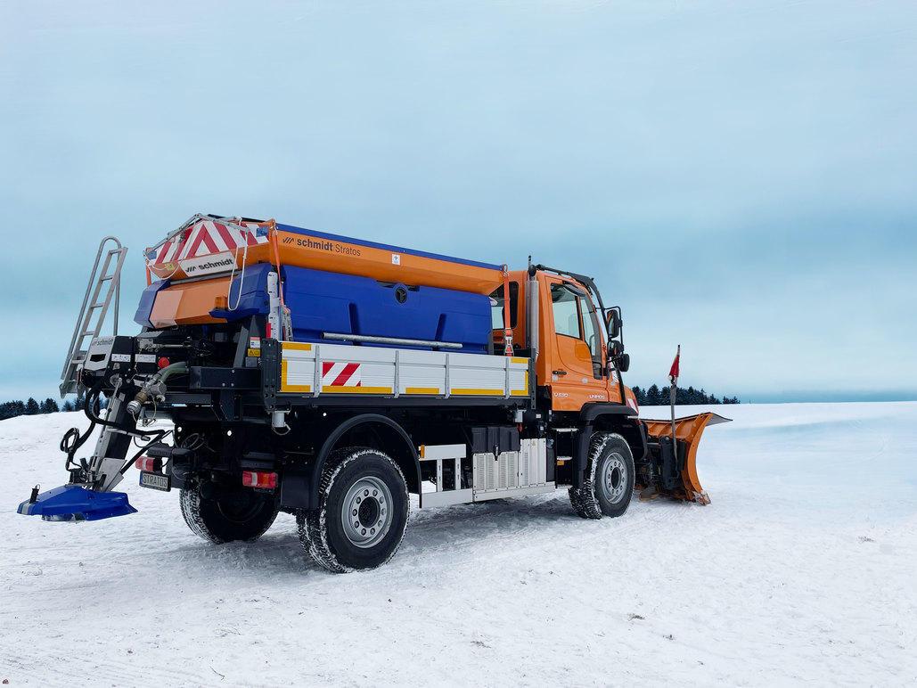 En snøplogbil står på snødekt vei med blå og oransje farger, klar til å rydde snø.