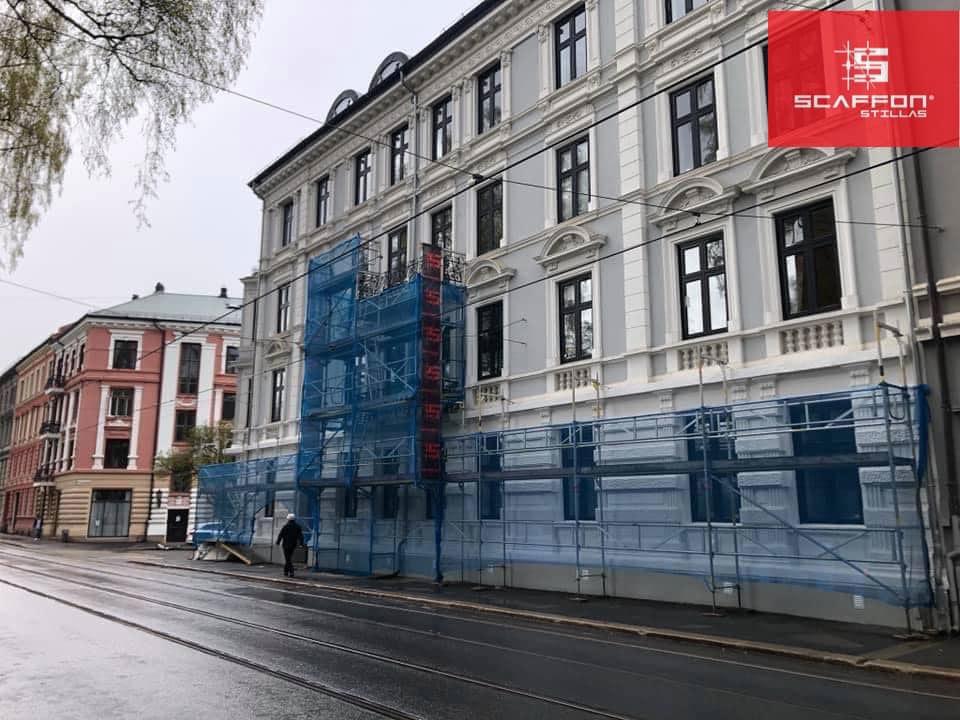 Bygning under rehabilitering med stillas og blå nett, beliggende langs en gate i byen.