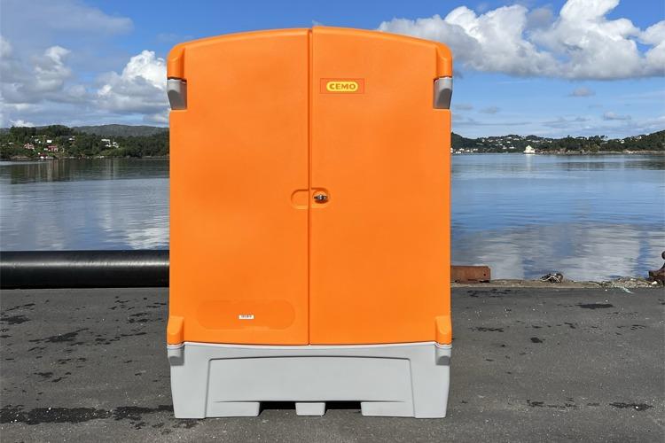En oransje toalettkabin med grå bunn, plassert ved vannkanten under en blå himmel.