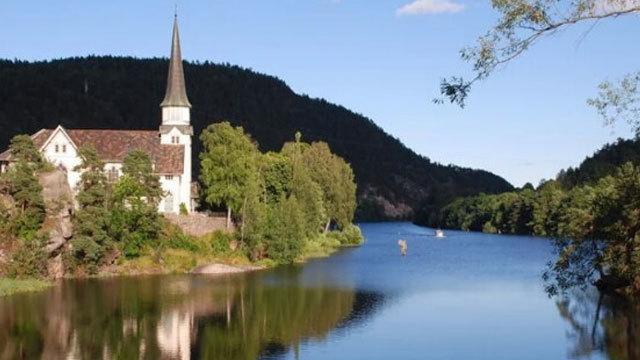 En vakker kirke ved elven med grønne trær og fjell i bakgrunnen under en klar blå himmel.
