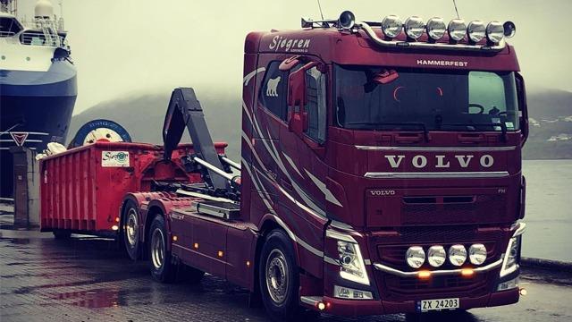En rød Volvo lastebil med tilhenger står ved havnen, med en stor container bak. Det er grått vær.