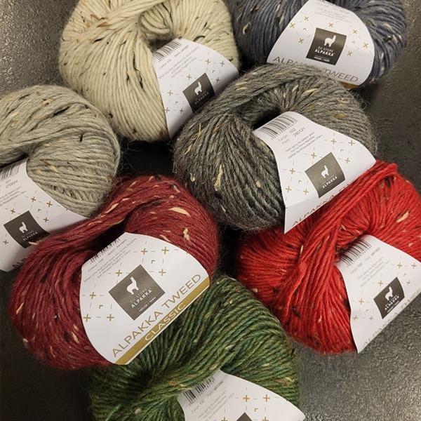 Fargerike garnnøster i alpakka tweed, inkludert rød, grønn, grå og beige, stablet på en overflate.