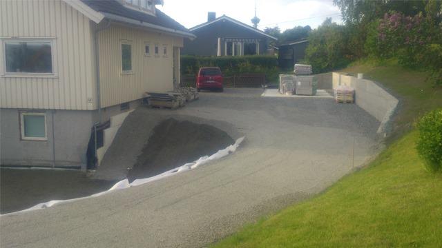 En gruslagt innkjørsel ved en bolig med en rød bil parkert, omgitt av grønt gress og trær.