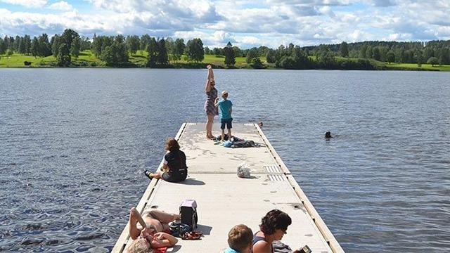 Familie som nyter en solfylt dag på en brygge ved innsjøen, med barn som leker og voksne som slapper av.