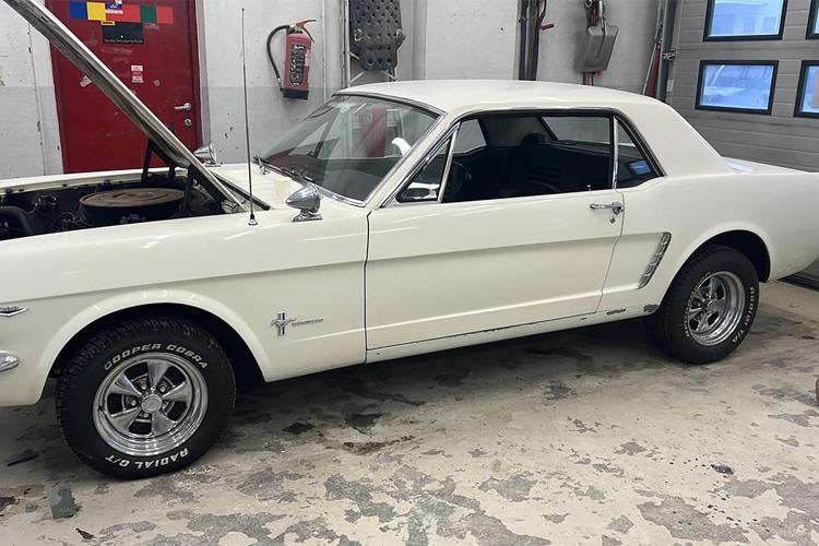 En hvit 1964 Ford Mustang med åpen motorlokk, parkert i en verkstedlignende setting.