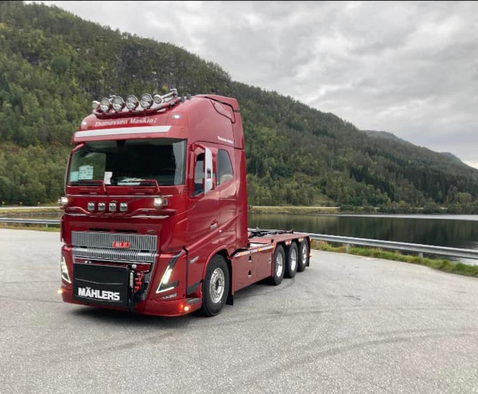 Rød lastebil med høy kabin parkert ved en innsjø, omringet av grønne fjell.