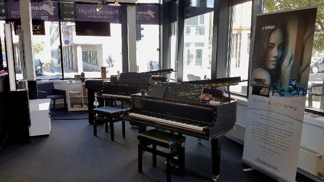 To pianoer i en butikk med store vinduer, stoler og en plakat med en kvinne i bakgrunnen.