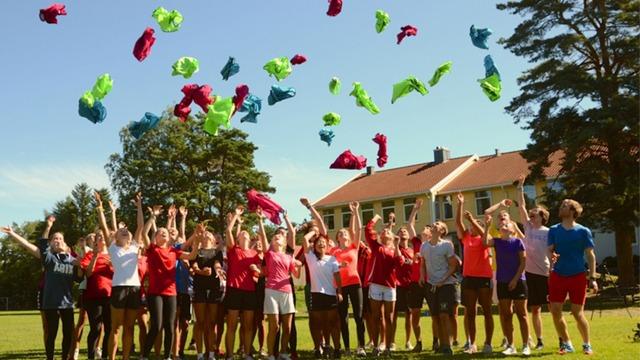 Gruppe ungdommer kaster fargerike klær i luften på en solfylt dag på en skolegård.