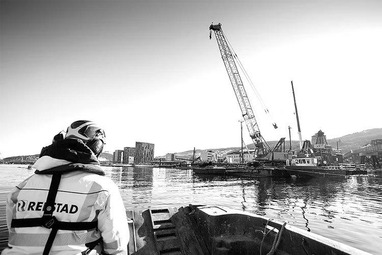 En person i båt ser på en kran som arbeider ved havnen i en by. Bygninger og fjell i bakgrunnen.