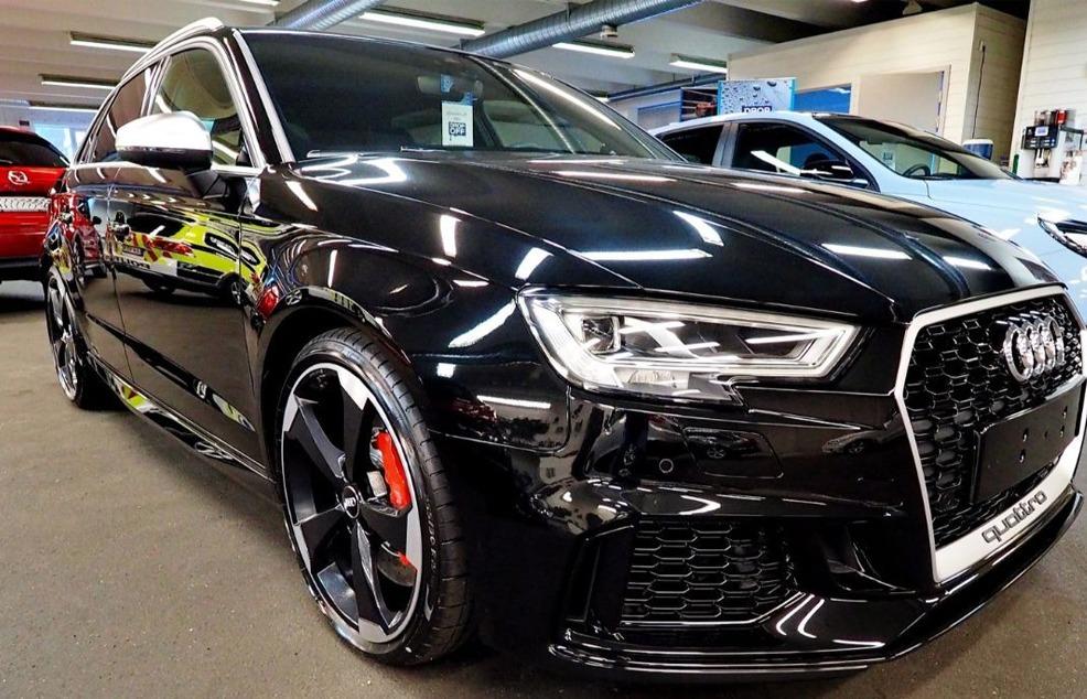 En svart Audi RS3 med sportslige detaljer, stående i en bilutstilling med andre biler i bakgrunnen.