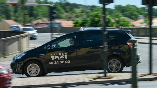 En svart taxi med teksten "Halden Taxi" kjører på en vei i Halden.