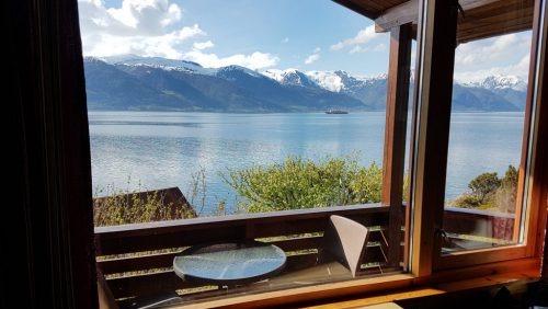 Utsikt fra et vindu med fjell og innsjø, med en terrasse med bord og stoler i forgrunnen.