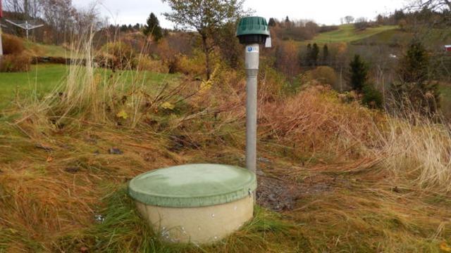 En grønn brønn med lokk på en hage, omgitt av gress og busker i et naturskjønt område.