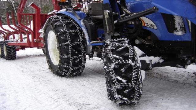 Traktor med snøkjettinger på dekkene, parkert på en snødekt vei ved siden av en redskapstralle.