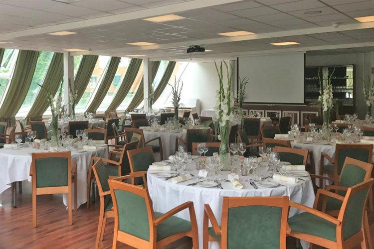 Elegant restaurant med dekkede bord, blomsterdekorasjoner og store vinduer som slipper inn naturlig lys.