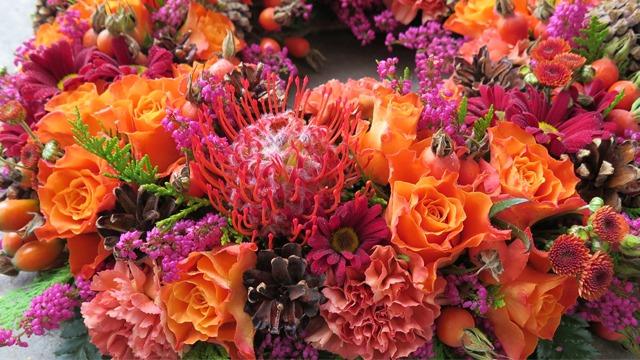 En fargerik blomsterkrans med oransje roser, lilla krysantemum og røde protea-blomster.