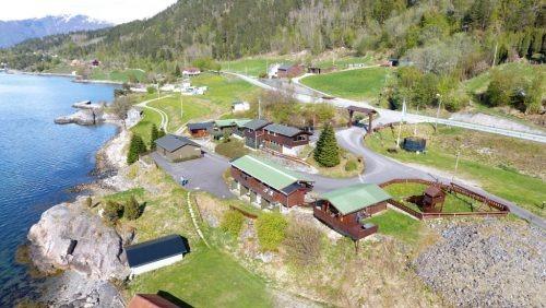 Luftbilde av en kystlandsby med hytter, veier og grøntområder ved sjøen. Fjell i bakgrunnen.