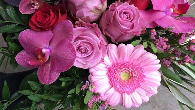 En fargerik bukett med roser, orkideer og gerbera i nyanser av rosa og lilla, omkranset av grønt bladverk.