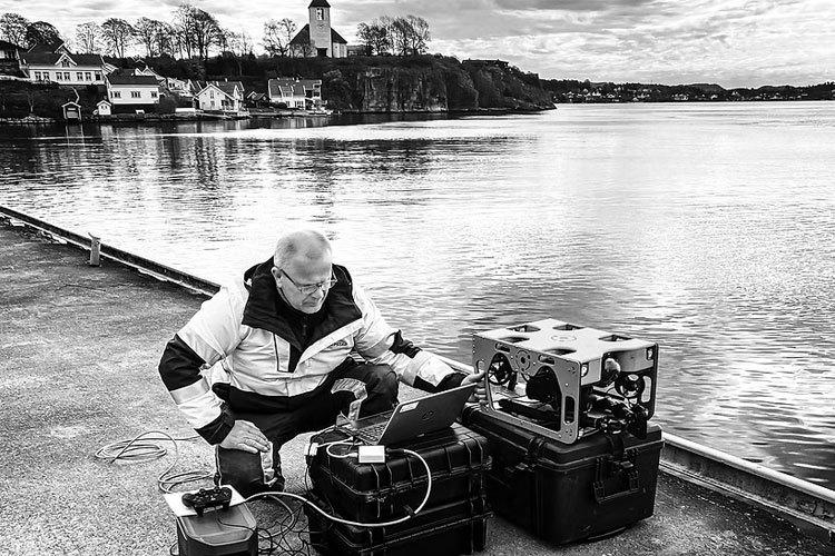 En mann sitter på kaien og bruker en bærbar datamaskin med utstyr ved vannet.