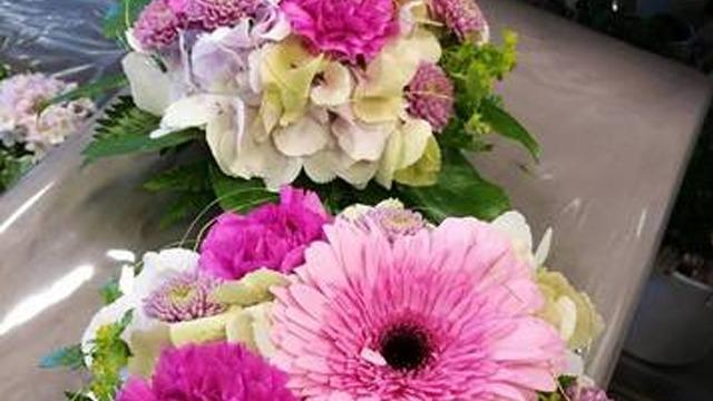To vakre blomsteroppsatser med rosa og lilla blomster, inkludert gerbera og hortensia, på en lys bakgrunn.