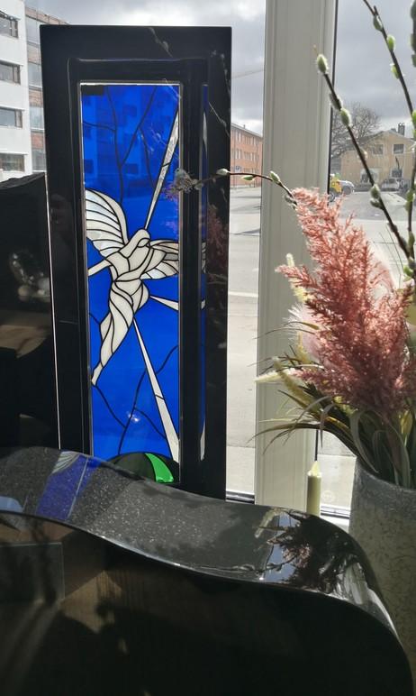Farget glassvindu med engelmotiv, plassert ved et vindu med blomster i forgrunnen.