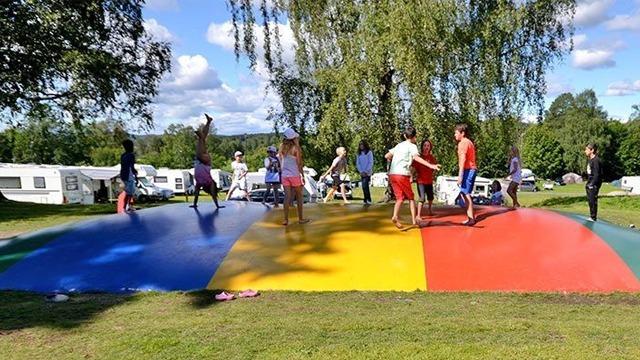 Barn og unge hopper på en stor fargerik hoppematte i en campingplass med trær i bakgrunnen.