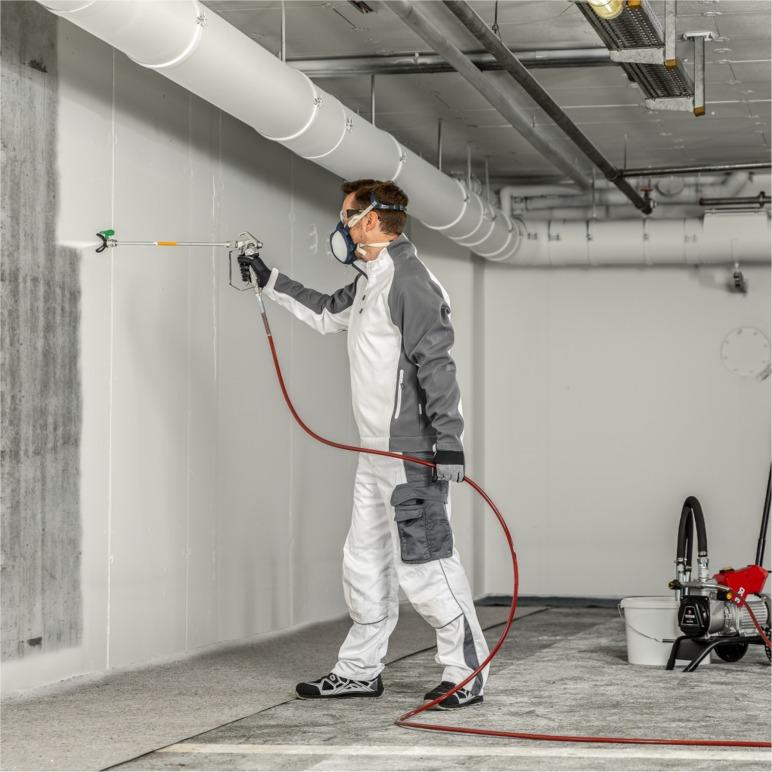 En maler i hvit arbeidsdrakt sprayer veggen i et rom med grå betong.