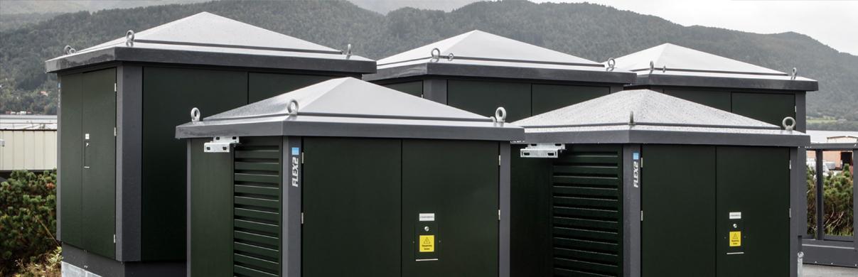 Fire grønne transformatorbokser med skråtak plassert på en plattform, med fjell i bakgrunnen.