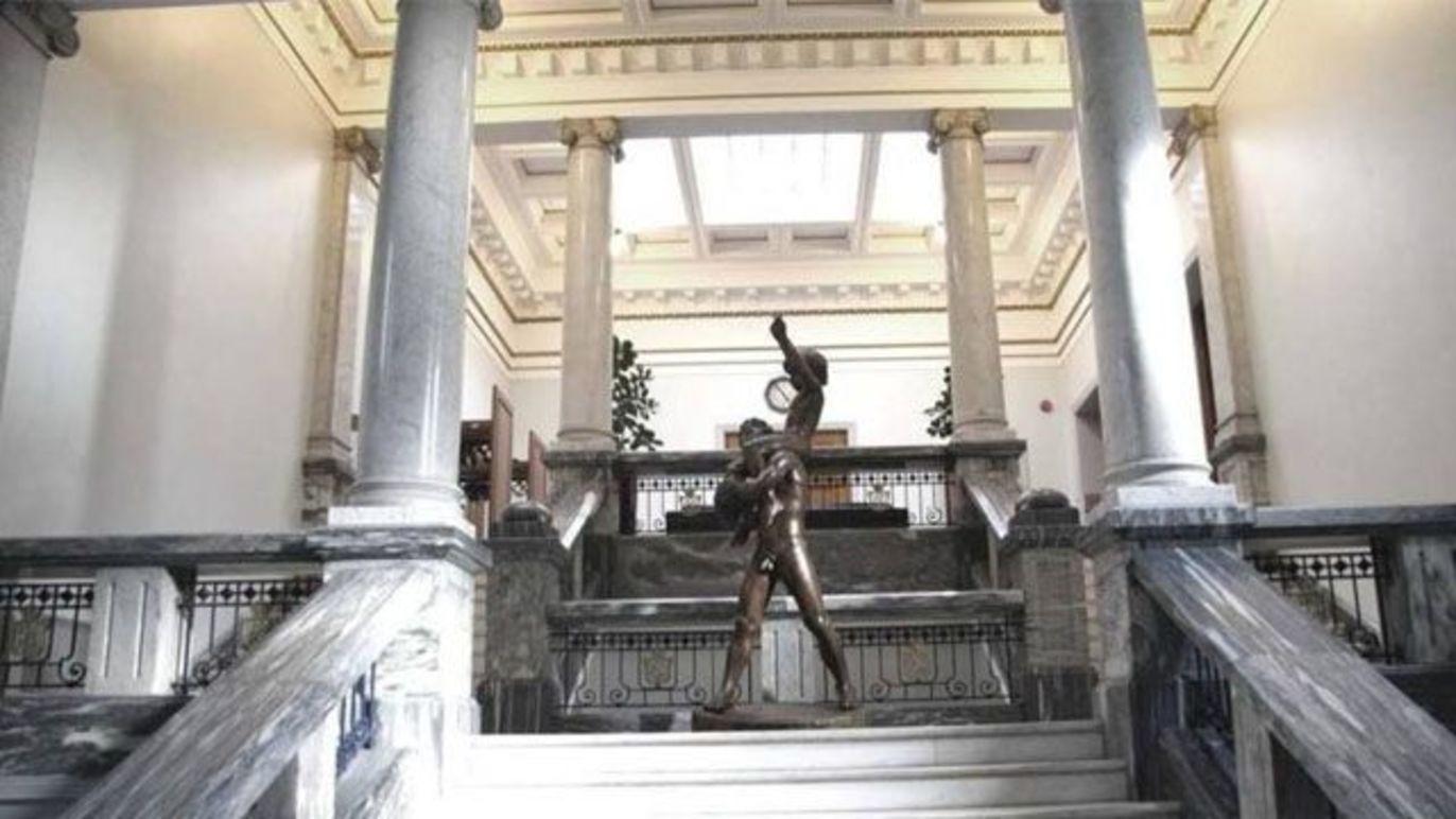 En skulptur av en kriger står i en stor hall med søyler og naturlig lys som strømmer inn fra taket.