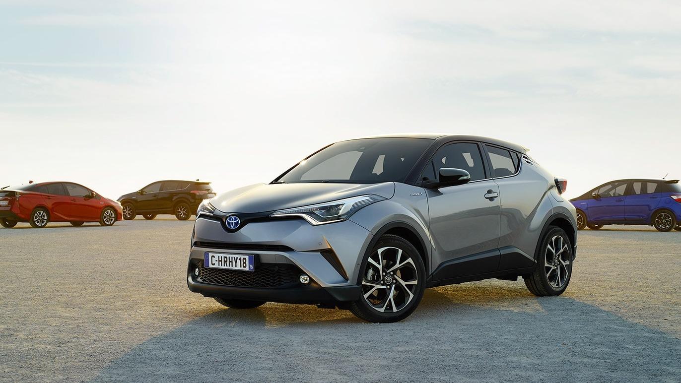 En sølvfarget Toyota C-HR står på en sandaktig overflate med flere biler i bakgrunnen.