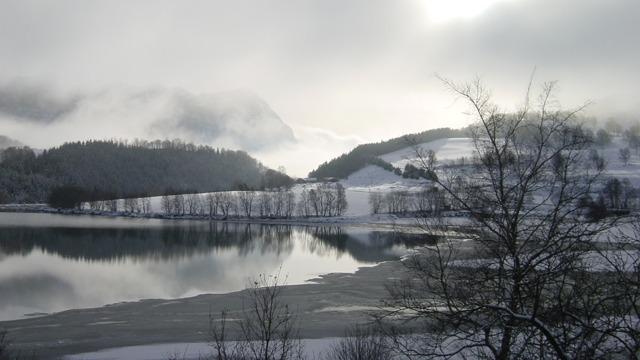 Vinterlandskap med snødekte fjell, en rolig innsjø og tåke som svever over landskapet.