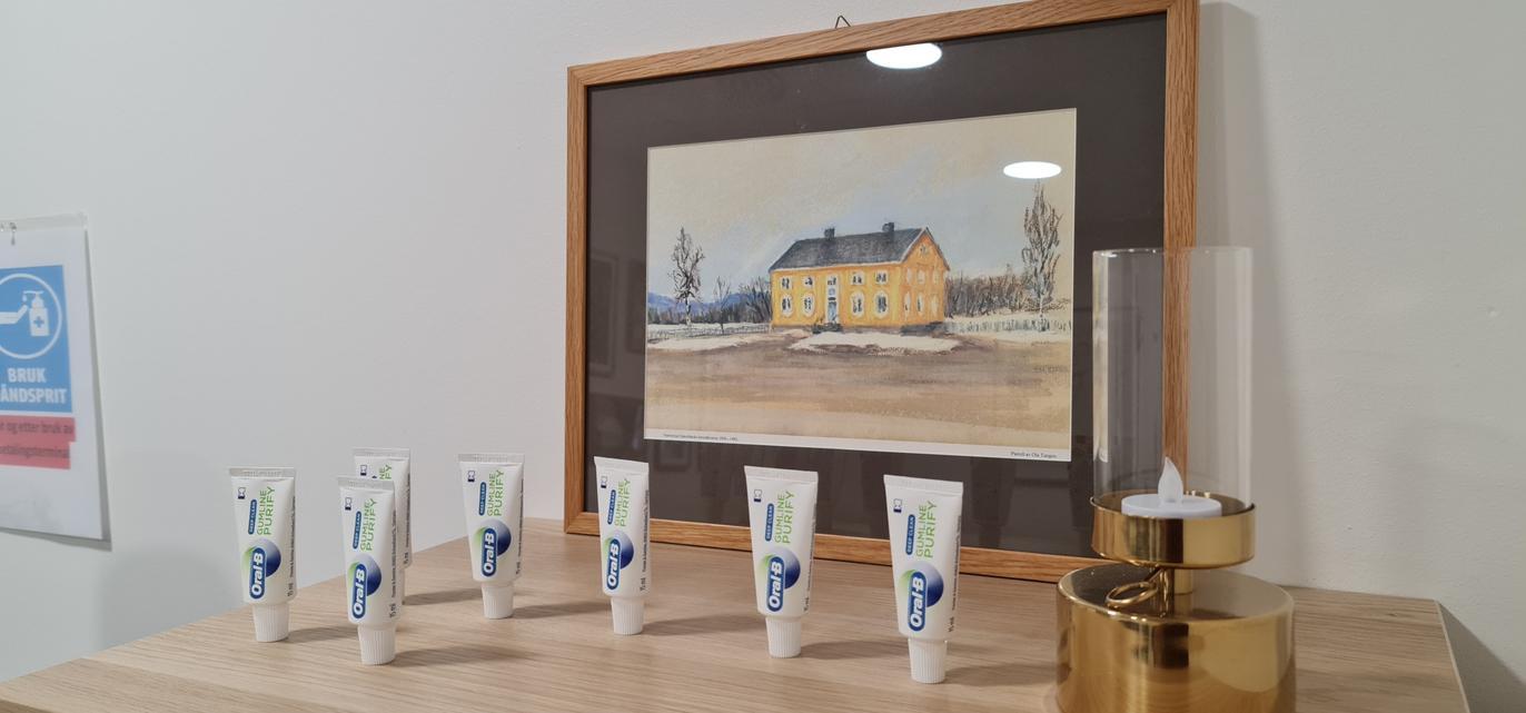 En rammet akvarell av et gult hus med snø, med seks kremtuber og en gullfarget beholder foran.