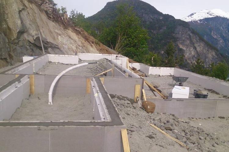 Byggeplass med fundament for hus, med fjell og snødekte topper i bakgrunnen.