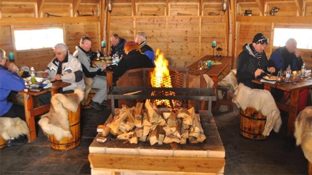 Innendørs restaurant med folk som spiser rundt en stor peis, omgitt av trevegger og koselige sitteplasser.