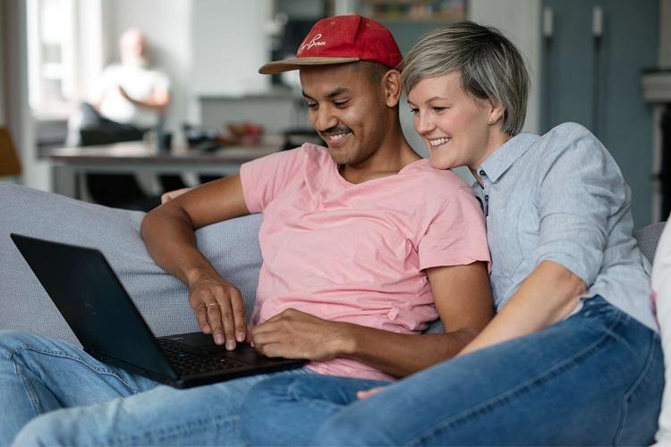 To personer sitter sammen på en sofa og ser på en bærbar datamaskin. De smiler og ser glade ut.