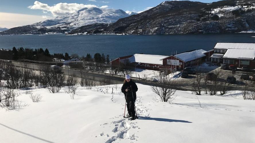 En person går i snøen med fjell og en innsjø i bakgrunnen, omringet av snødekte trær.