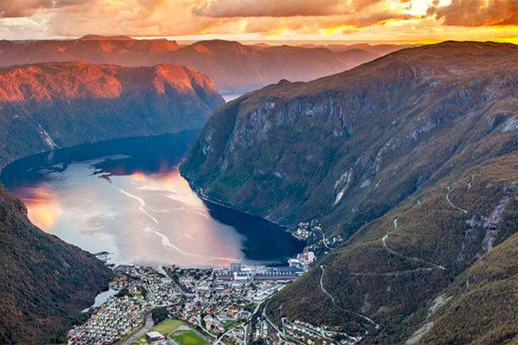 En panoramautsikt over en fjord med fjell og en liten by ved vannkanten under solnedgang.