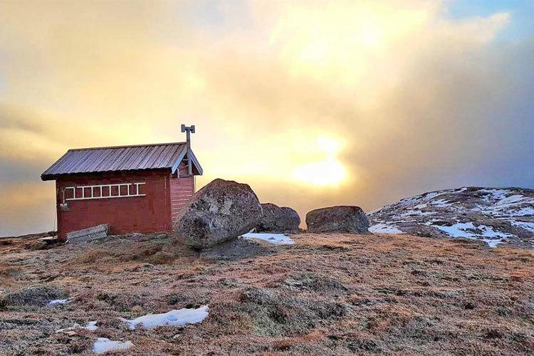 Rød hytte på fjellet med steiner og snø, mot en solfylt himmel med skyer i bakgrunnen.