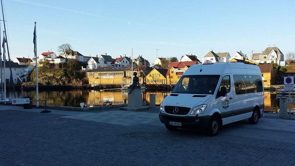 En hvit varebil står ved havnen med bygninger og båter i bakgrunnen under en klar blå himmel.
