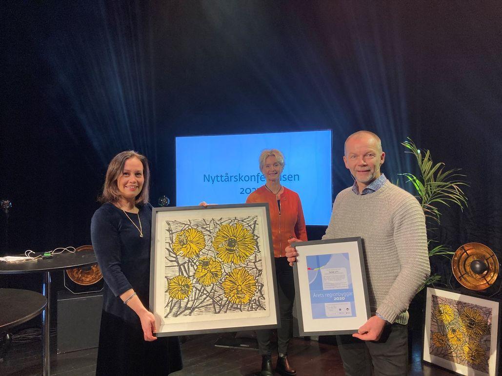 Tre personer står på en scene med kunstverk og en pris, med en skjerm i bakgrunnen som viser teksten "Lyndalensprisen 2023".