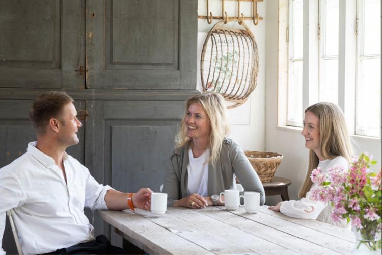 Tre personer sitter rundt et bord i et lyst rom, nyter kaffe og samtaler, med blomster og kurver i bakgrunnen.