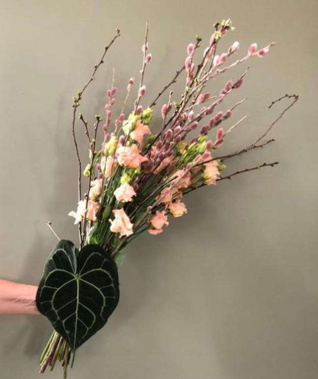 En bukett med blomster og grener, inkludert rosa og hvite blomster, holdt av en hånd mot en grå bakgrunn.
