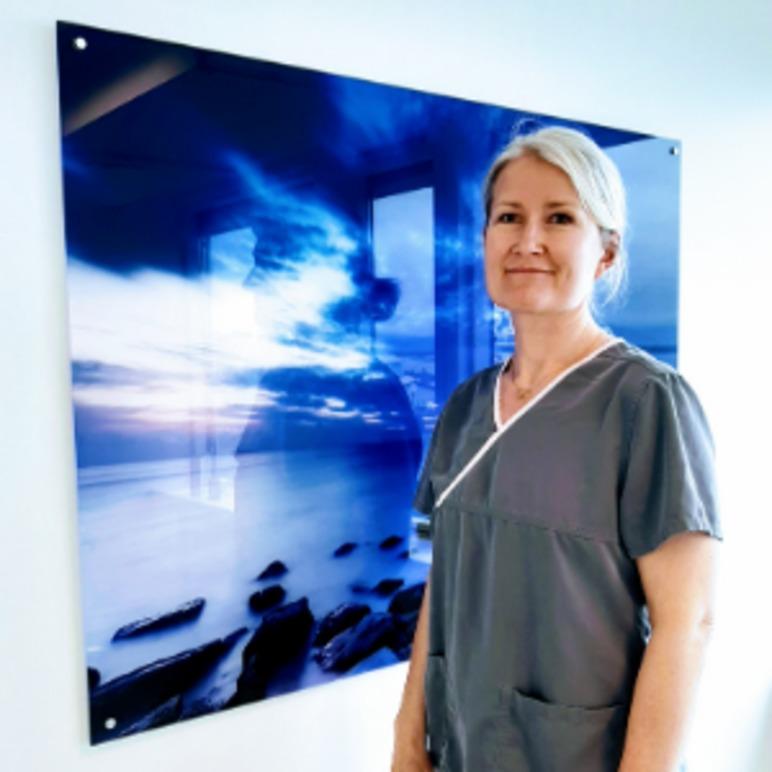 En kvinne i grå arbeidsdrakt står foran et stort bilde av en havutsikt med steiner og dramatisk himmel.