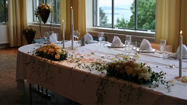 Et elegant festbord dekket med hvit duk, blomsterdekorasjoner og stearinlys, med utsikt over vannet.