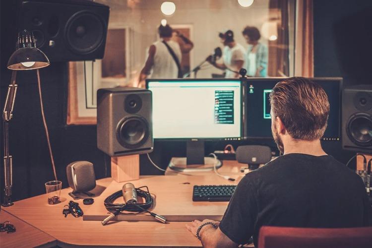 En person sitter ved et skrivebord med datamaskiner og høyttalere, mens musikere øver i bakgrunnen.