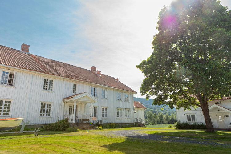 Et stort, hvitt hus med rød tak og grønne plener, omgitt av trær og fjell i bakgrunnen.