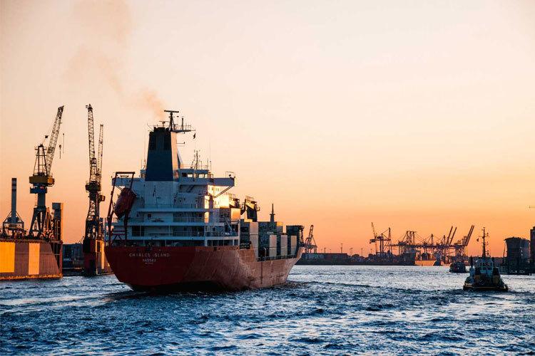 Et fraktskip seiler ut av havnen ved solnedgang, med kraner og andre skip i bakgrunnen.