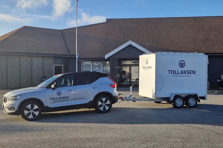 En bil med tilhenger fra Tollaksen står parkert foran en bygning med tak i brune farger.