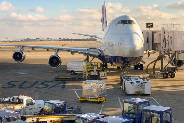 Et stort passasjerfly står ved en flyplassterminal med bagasjevogner og lasteutstyr rundt.