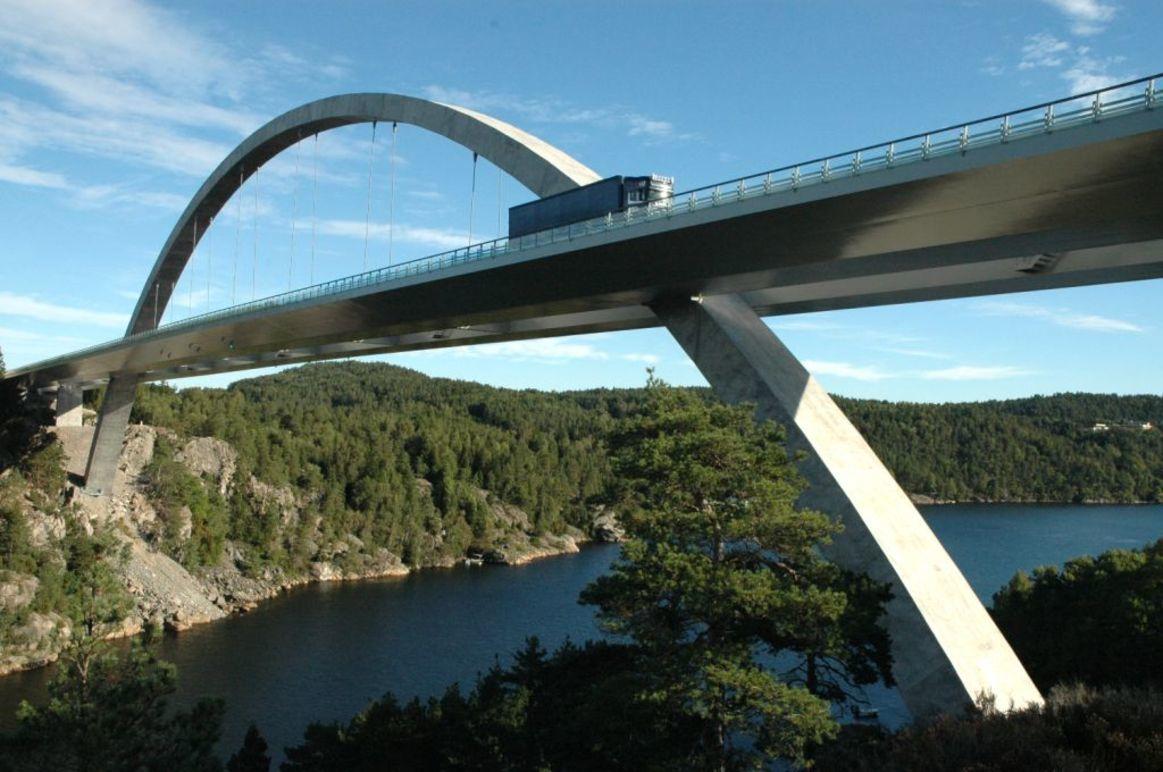 En moderne bro med en buet struktur som går over en innsjø, omgitt av skogkledde fjell.